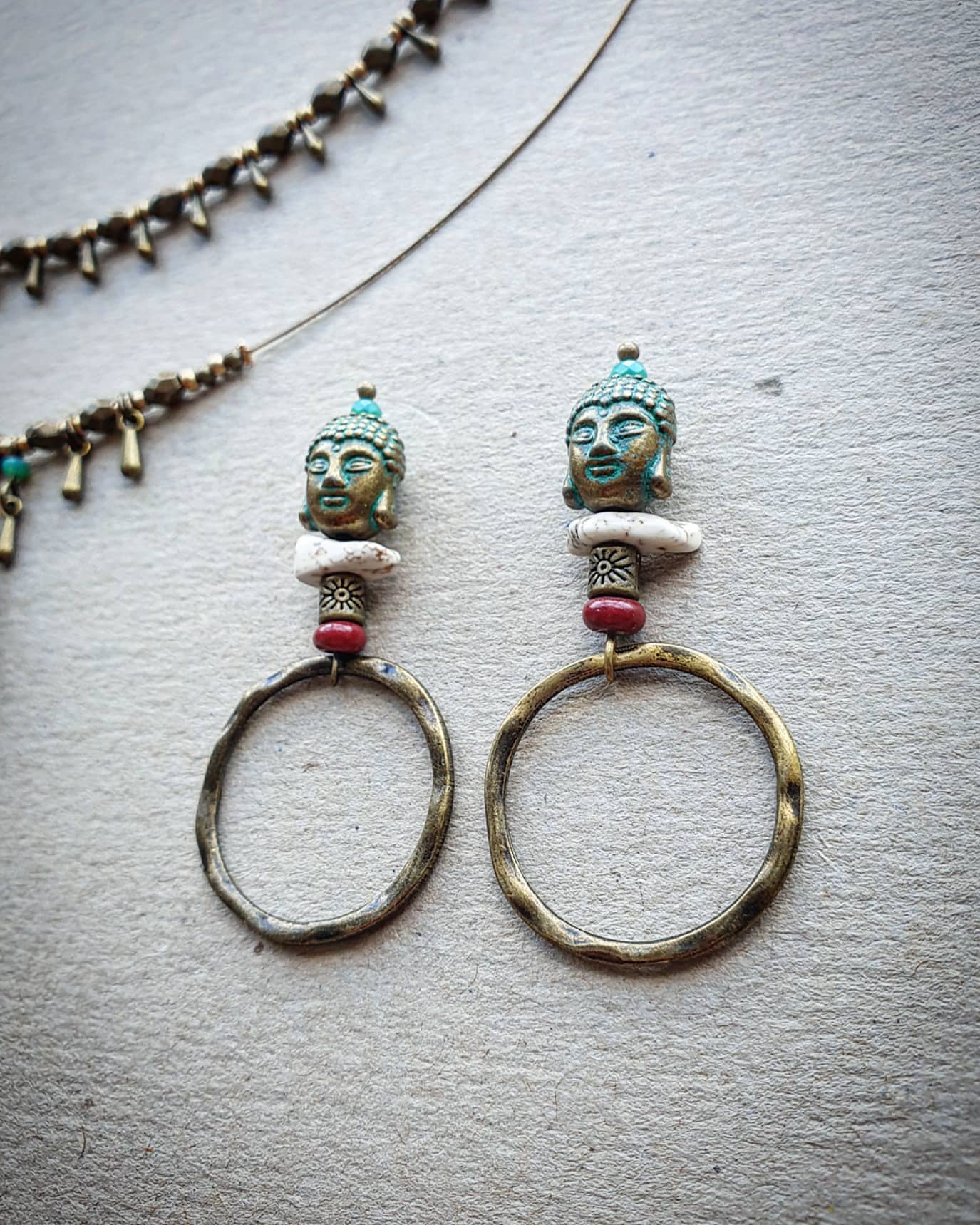 Pair of earrings with Buddha head designs and red and white beads on a light gray background