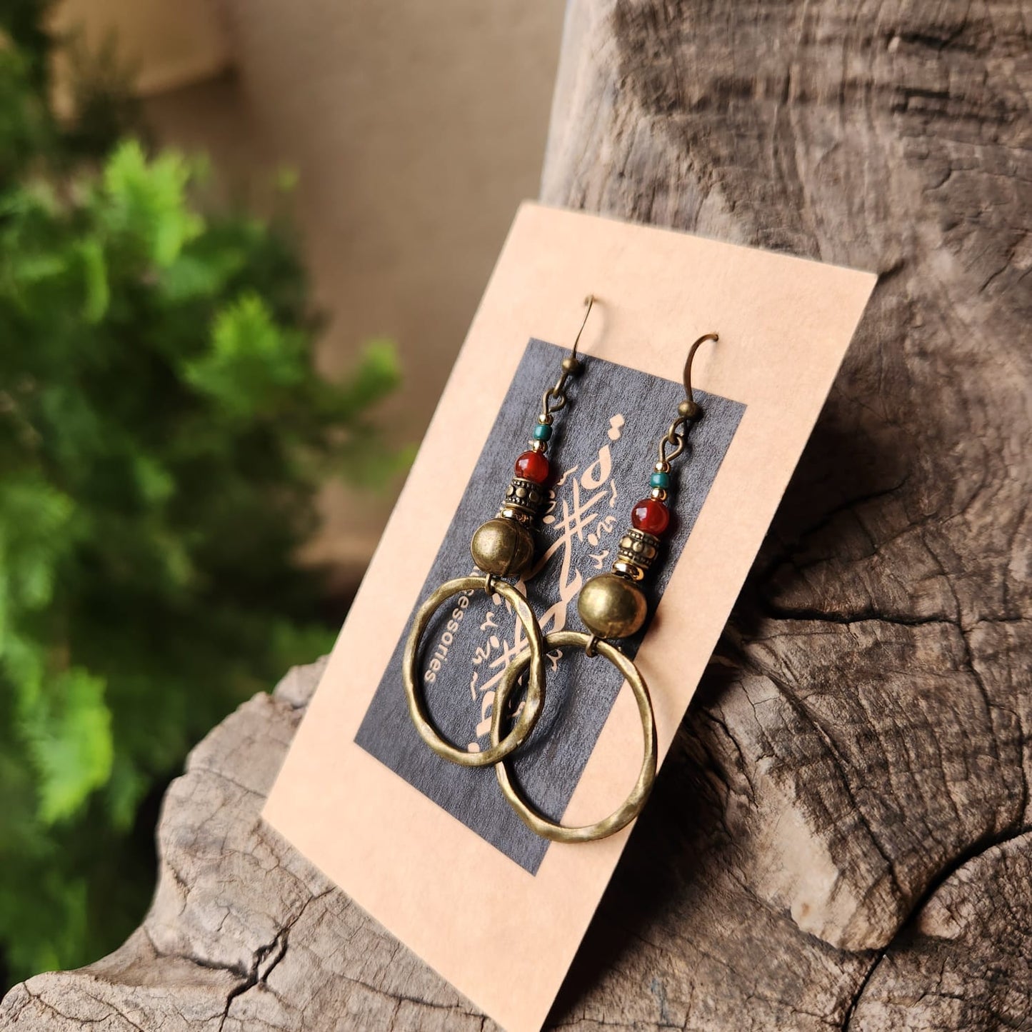 Pair of bronze earrings with colorful beads on a card, placed on a wooden surface with greenery in the background.