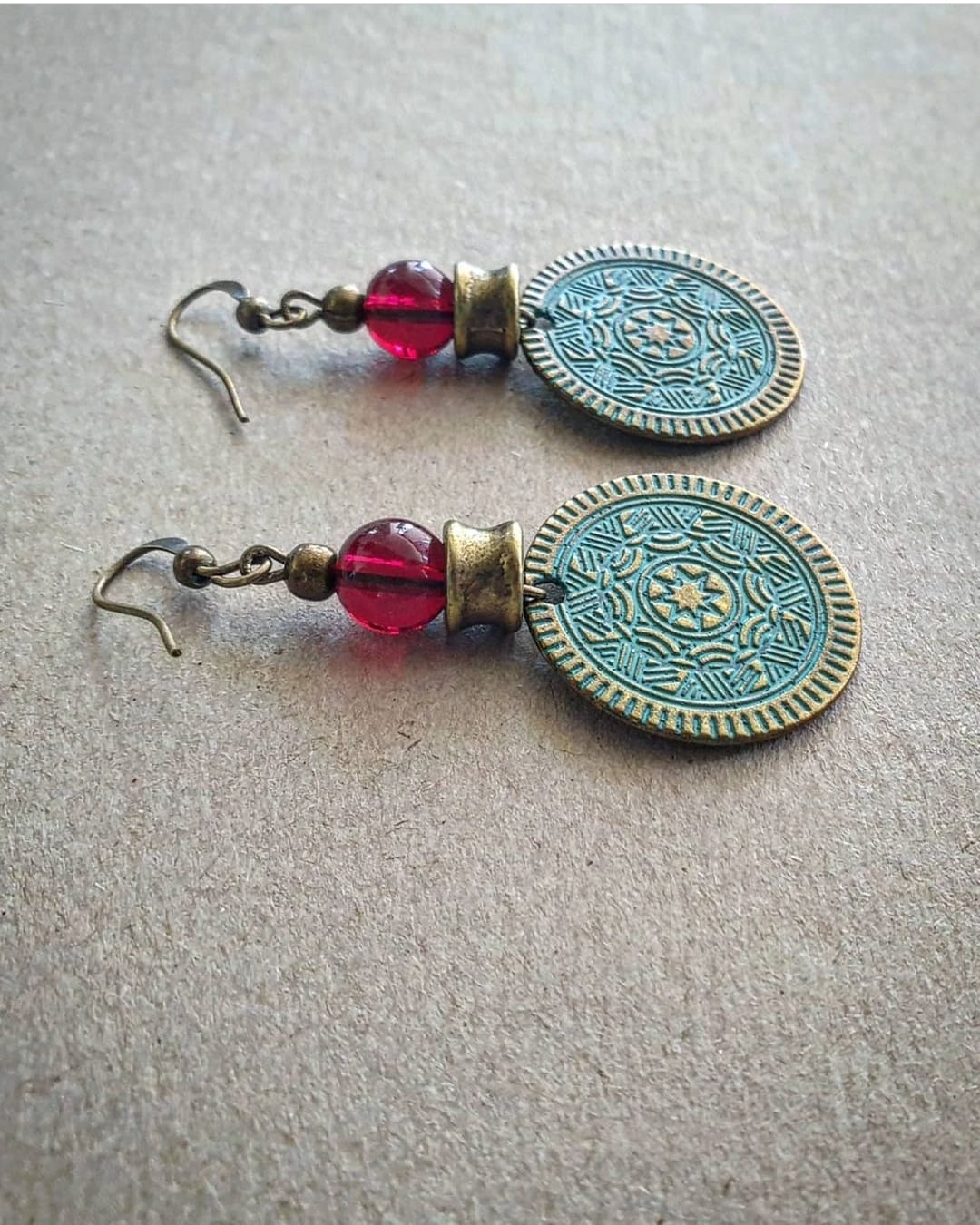 A pair of bronze coin earrings with intricate designs and red beads attached to them.
