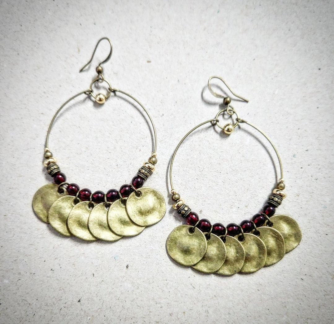 A close-up of a bohemian-style brass hoop earring. The antique brass hoop is decorated with deep-red beads and several hanging, hammered brass coin charms.