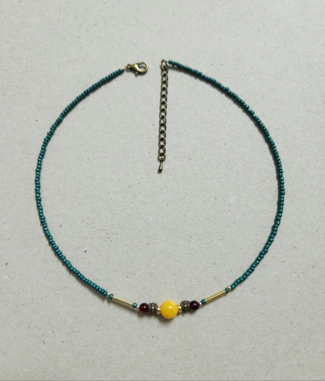 A delicate, handmade beaded necklace consists of small teal seed beads with a central focal arrangement featuring a large yellow bead flanked by gold plated brass spacers, deep-red beads, and gold-tone bar accents.