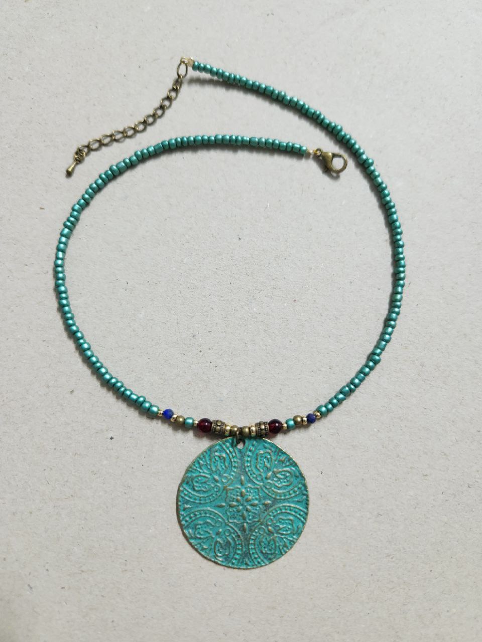 A bohemian-style necklace featuring a strand of shimmering teal beads. The centerpiece is a large, circular patina-green pendant attached to the strand with small red and gold plated accent beads.