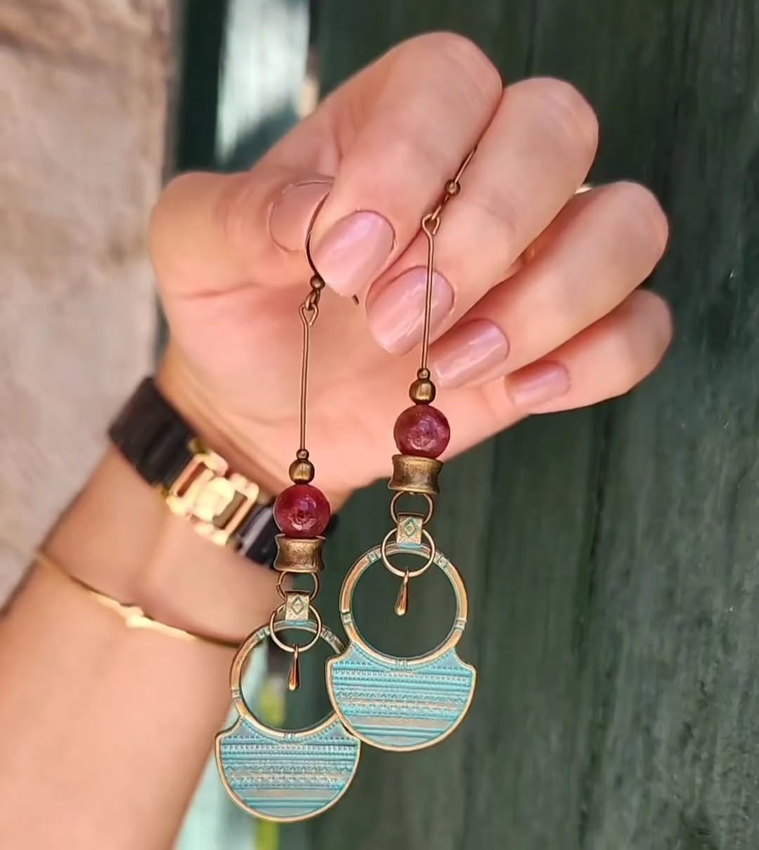 Hand holding a pair of bronze earrings with intricate designs in turquoise  and red