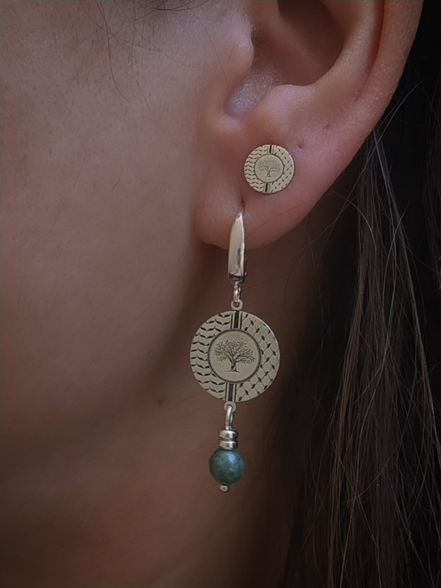 Close-up of an ear wearing a silver stud earring, underneath another silver drop earring with a green bead.