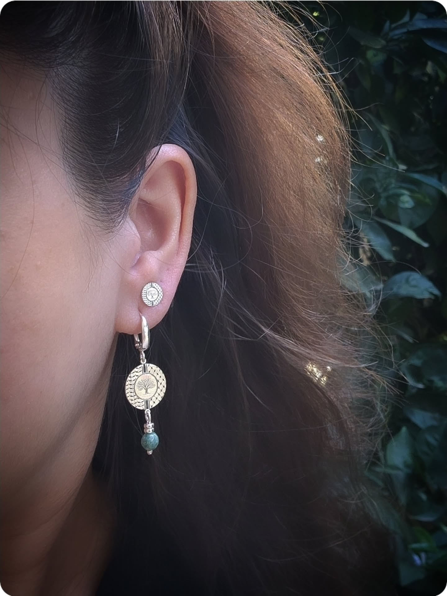 Close-up of an ear wearing a silver stud earring, underneath another silver drop earring with a green bead.