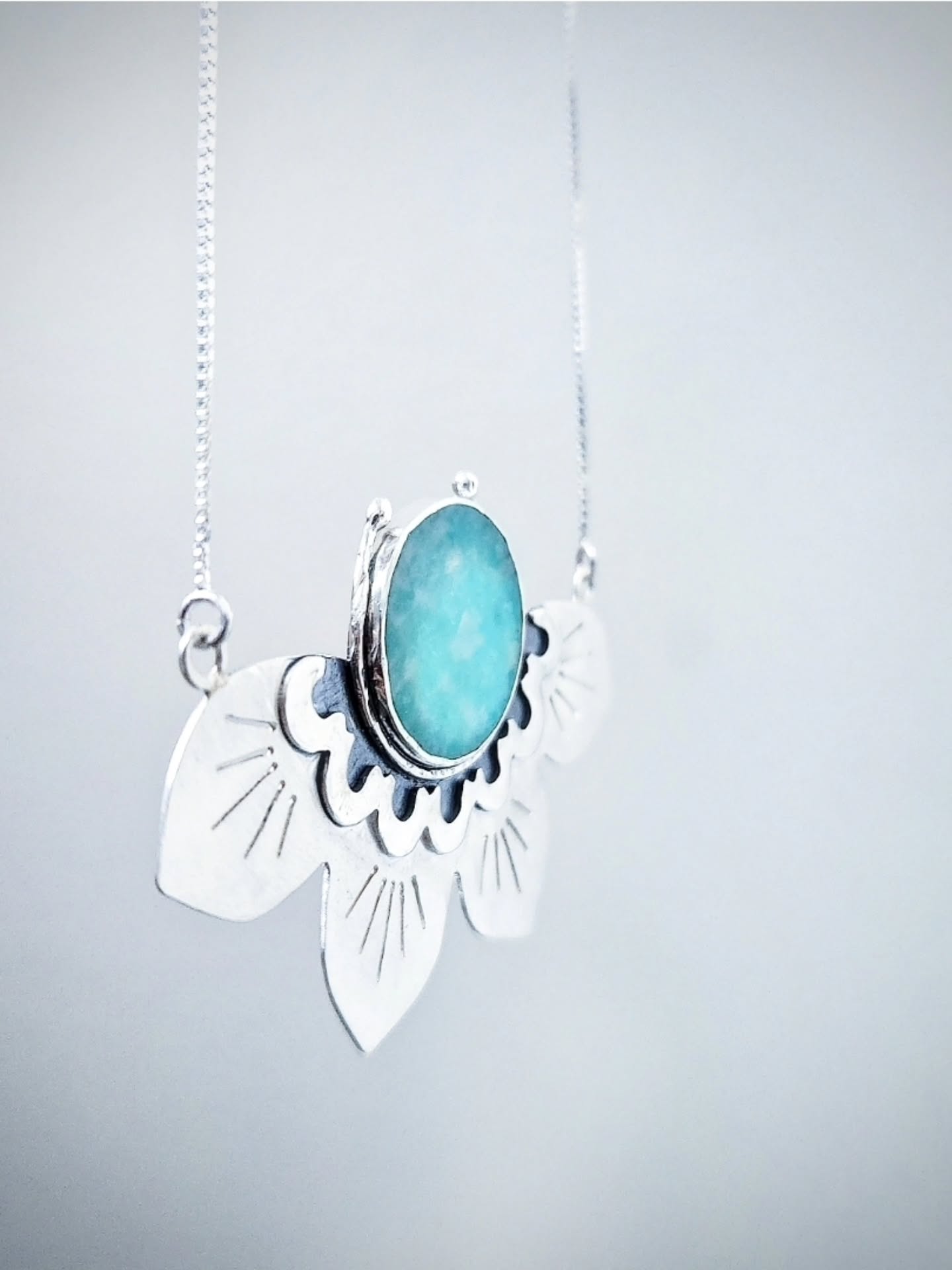 A person wearing a fine silver chain with a large half lotus-shaped pendant. The pendant features a central oval turquoise or amazonite stone set within silver petals decorated with engraved lines and blue enamel detailing.