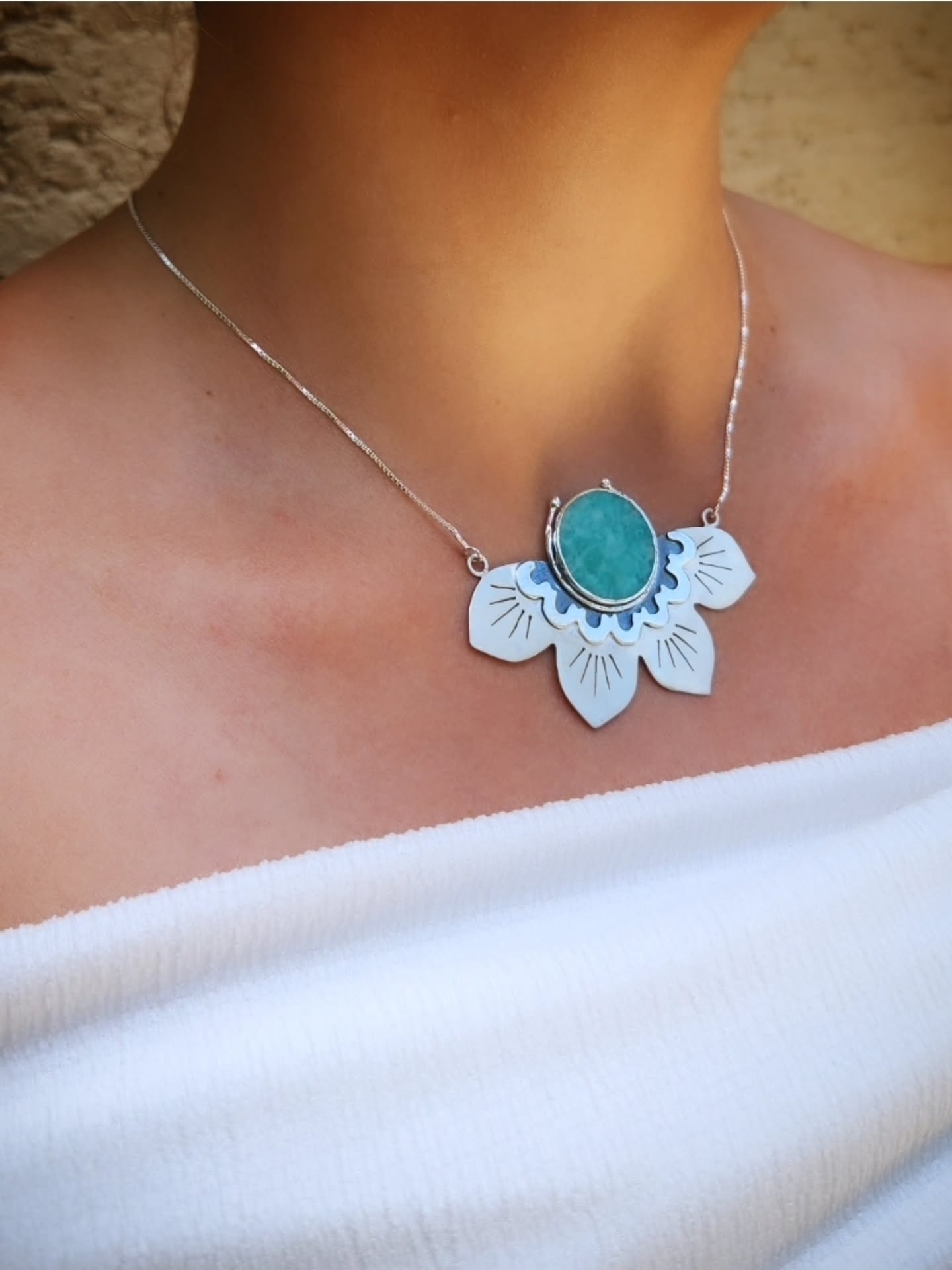 A person wearing a fine silver chain with a large half lotus-shaped pendant. The pendant features a central oval turquoise or amazonite stone set within silver petals decorated with engraved lines and blue enamel detailing.