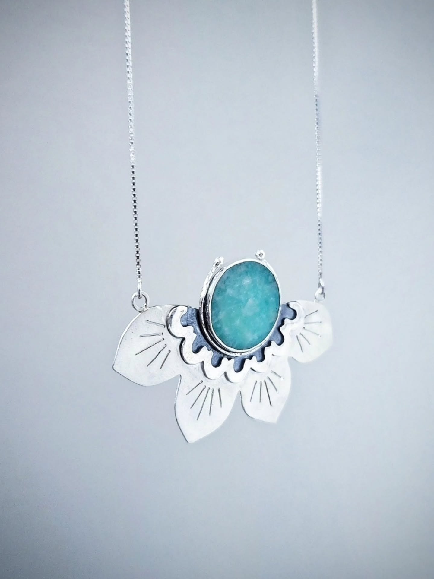 A person wearing a fine silver chain with a large half lotus-shaped pendant. The pendant features a central oval turquoise or amazonite stone set within silver petals decorated with engraved lines and blue enamel detailing.