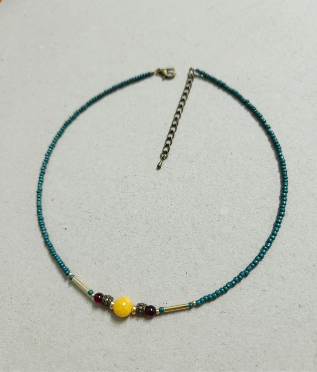 A delicate, handmade beaded necklace consists of small teal seed beads with a central focal arrangement featuring a large yellow bead flanked by gold plated brass spacers, deep-red beads, and gold-tone bar accents.