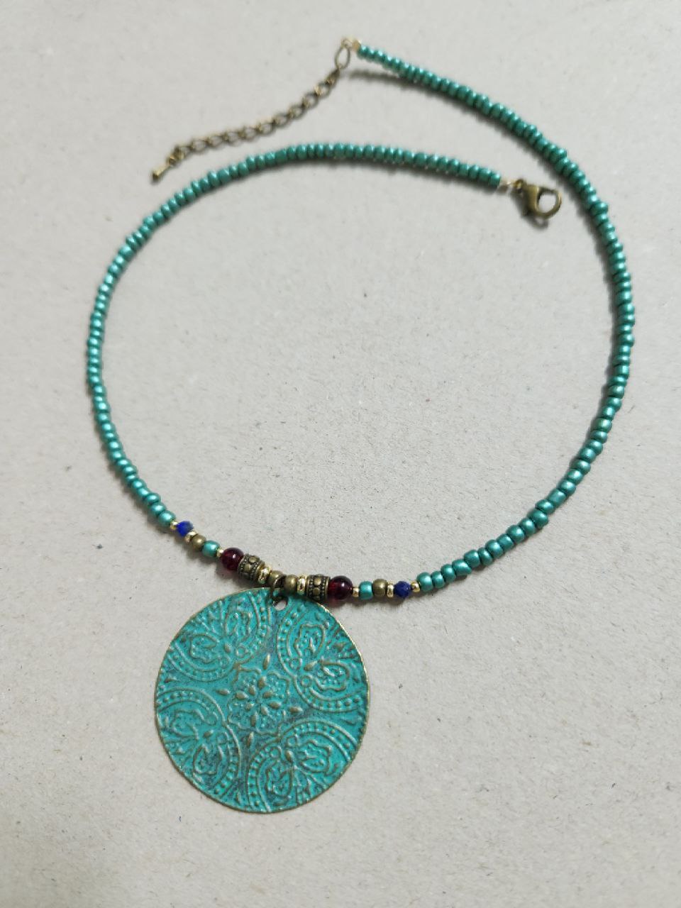 A bohemian-style necklace featuring a strand of shimmering teal beads. The centerpiece is a large, circular patina-green pendant attached to the strand with small red and gold plated accent beads.