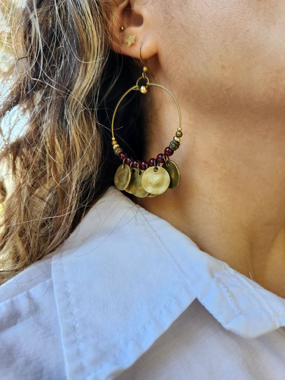 A close-up of a bohemian-style brass hoop earring. The antique brass hoop is decorated with deep-red beads and several hanging, hammered brass coin charms.