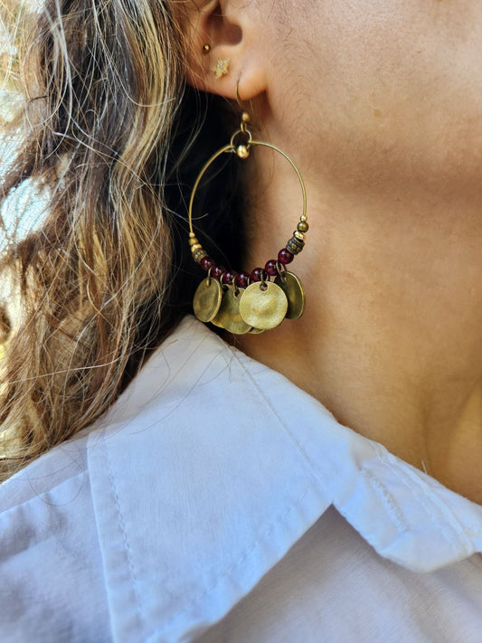 A close-up of a bohemian-style brass hoop earring. The antique brass hoop is decorated with deep-red beads and several hanging, hammered brass coin charms.