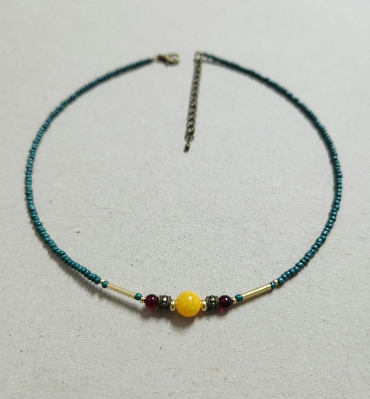 A delicate, handmade beaded necklace consists of small teal seed beads with a central focal arrangement featuring a large yellow bead flanked by gold plated brass spacers, deep-red beads, and gold-tone bar accents.