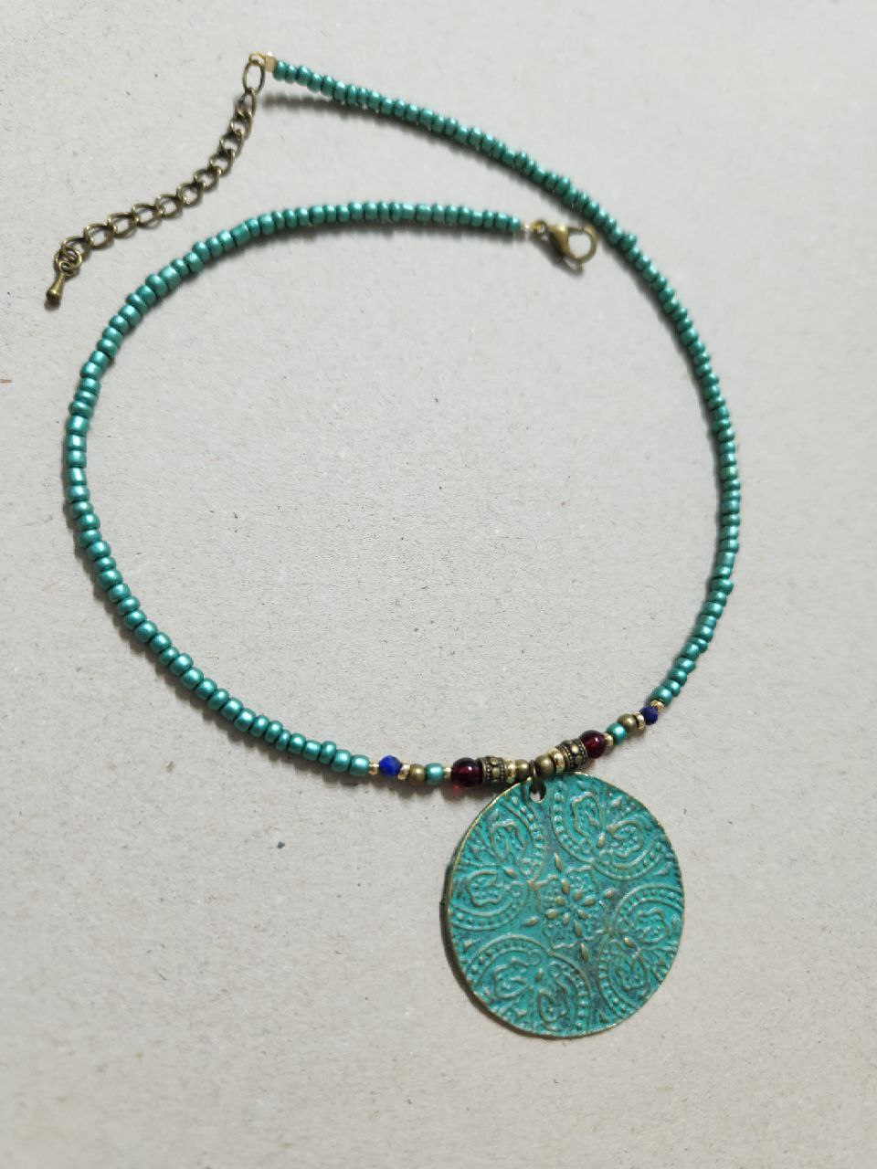 A bohemian-style necklace featuring a strand of shimmering teal beads. The centerpiece is a large, circular patina-green pendant attached to the strand with small red and gold plated accent beads.