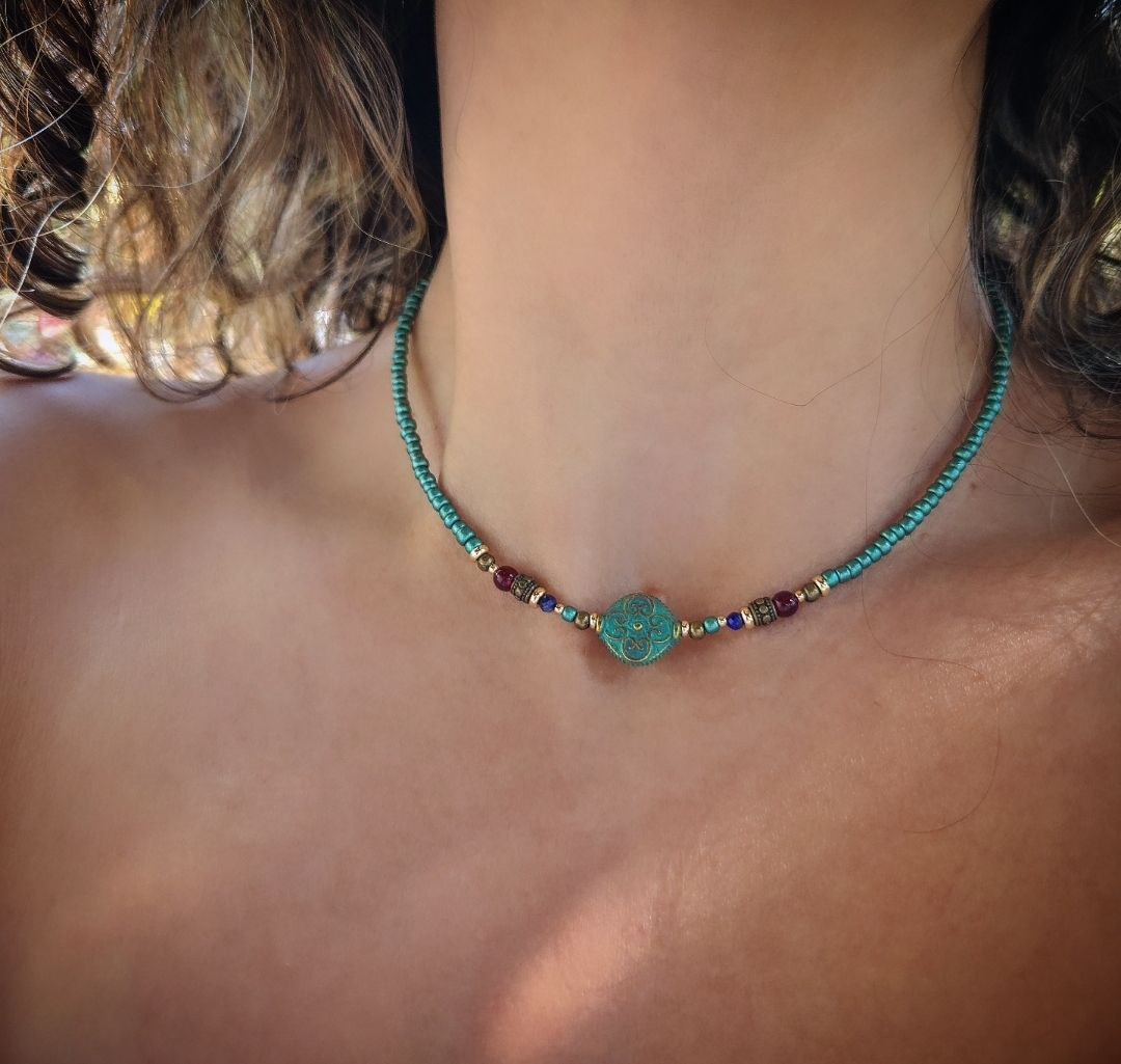 A handmade teal beaded necklace laid flat on a person's neck. The center features a round patina-green bead with an ornate scroll pattern, flanked by small red, blue, and gold brass decorative beads.