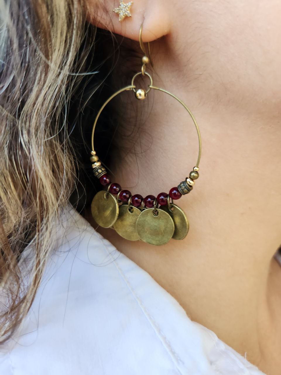 A close-up of a bohemian-style brass hoop earring. The antique brass hoop is decorated with deep-red beads and several hanging, hammered brass coin charms.