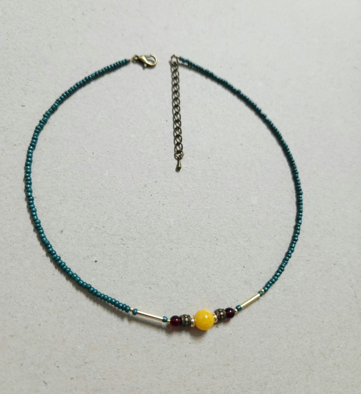 A delicate, handmade beaded necklace consists of small teal seed beads with a central focal arrangement featuring a large yellow bead flanked by gold plated brass spacers, deep-red beads, and gold-tone bar accents.