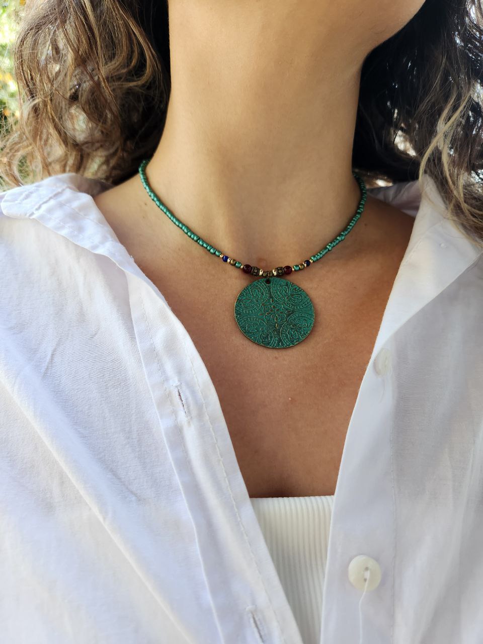 A person wearing a bohemian-style necklace featuring a strand of shimmering teal beads. The centerpiece is a large, circular patina-green pendant attached to the strand with small red and gold plated accent beads.