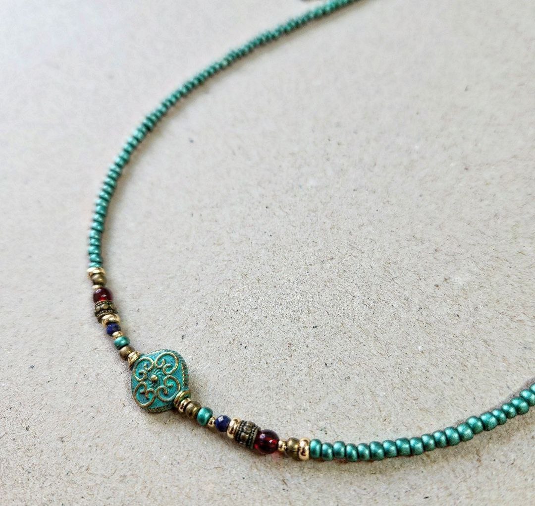 A handmade teal beaded necklace laid flat on a neutral surface. The center features a round patina-green bead with an ornate scroll pattern, flanked by small red, blue, and gold brass decorative beads.