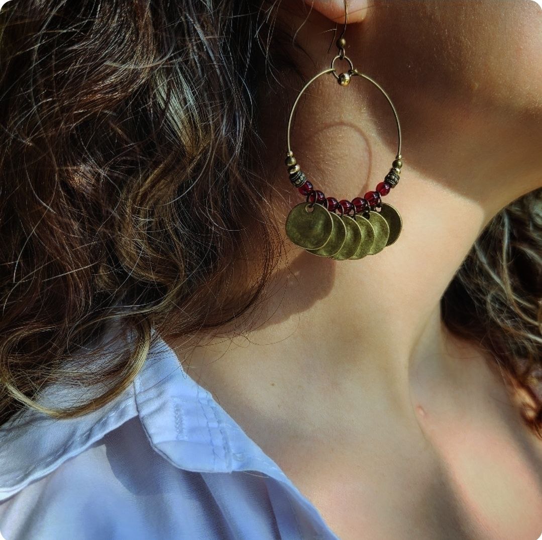 A close-up of a bohemian-style brass hoop earring. The antique brass hoop is decorated with deep-red beads and several hanging, hammered brass coin charms.