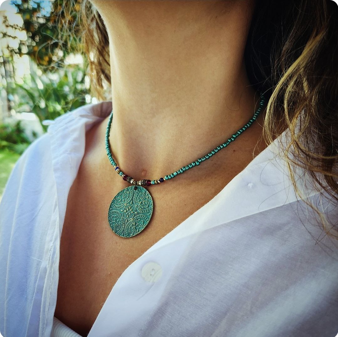 A person wearing a bohemian-style necklace featuring a strand of shimmering teal beads. The centerpiece is a large, circular patina-green pendant attached to the strand with small red and gold plated accent beads.