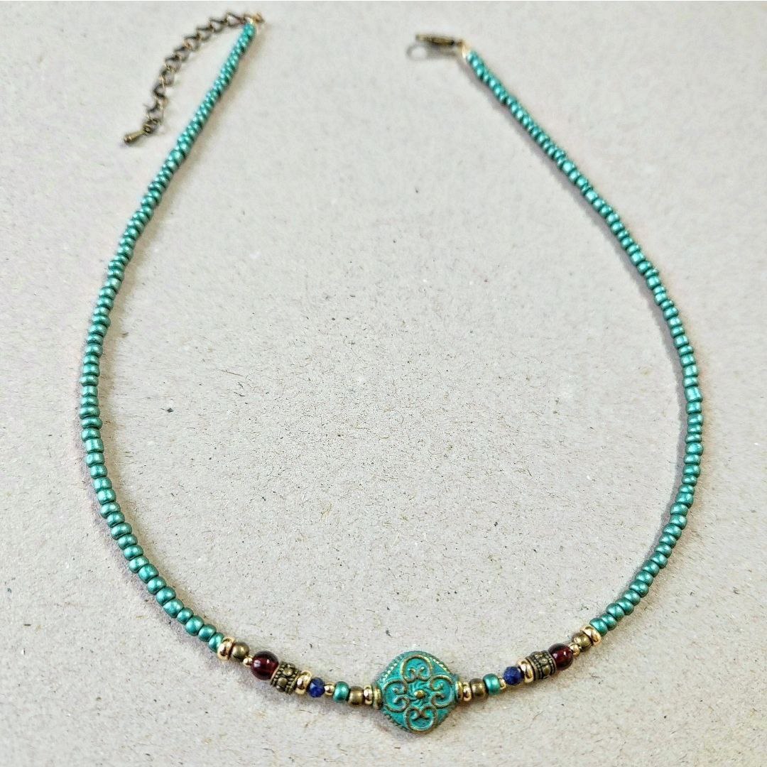 A handmade teal beaded necklace laid flat on a neutral surface. The center features a round patina-green bead with an ornate scroll pattern, flanked by small red, blue, and gold brass decorative beads.