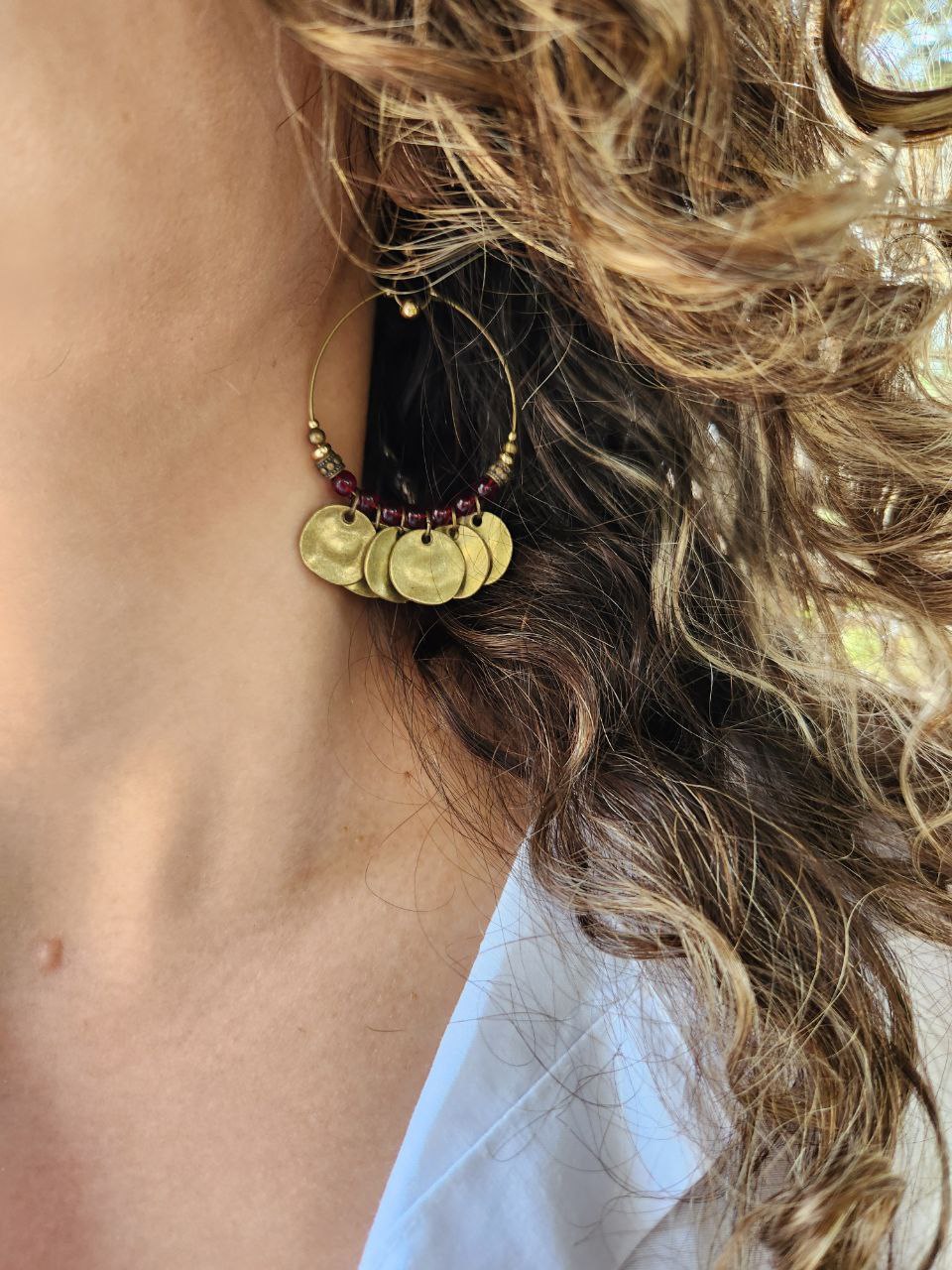 A close-up of a bohemian-style brass hoop earring. The antique brass hoop is decorated with deep-red beads and several hanging, hammered brass coin charms.