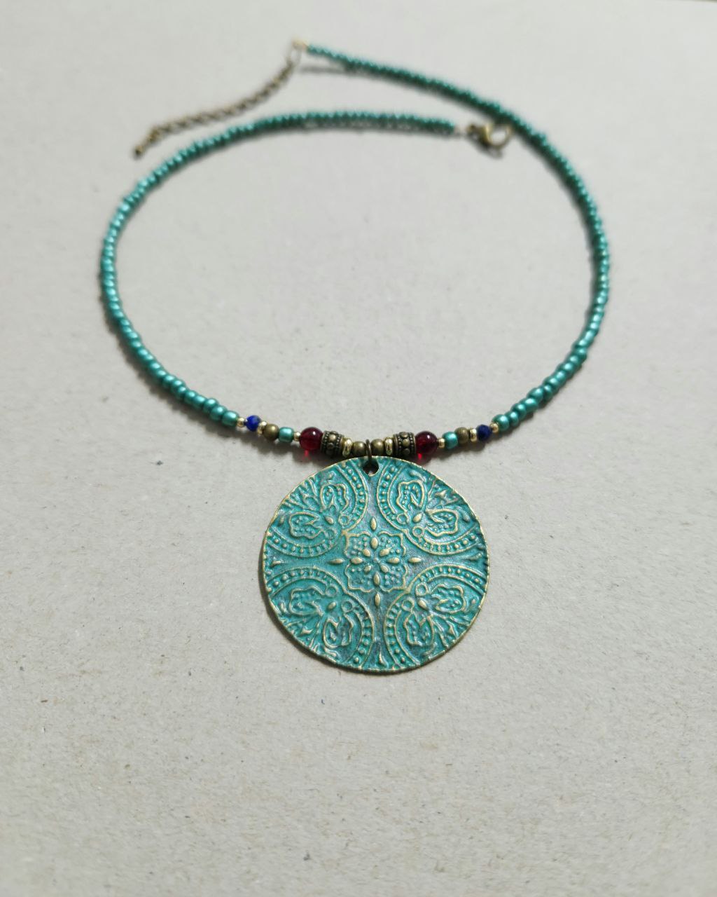 A bohemian-style necklace featuring a strand of shimmering teal beads. The centerpiece is a large, circular patina-green pendant attached to the strand with small red and gold plated accent beads.