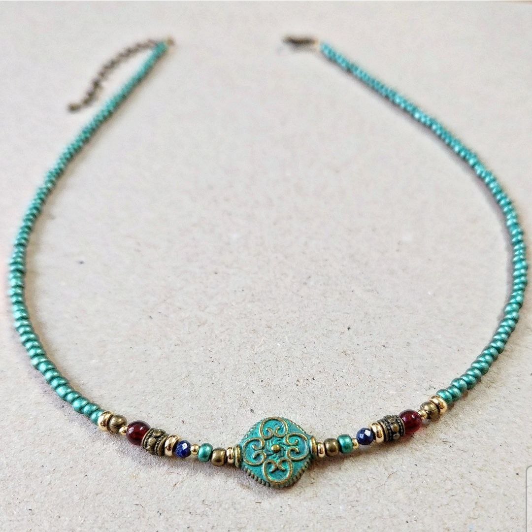 A handmade teal beaded necklace laid flat on a neutral surface. The center features a round patina-green bead with an ornate scroll pattern, flanked by small red, blue, and gold brass decorative beads.