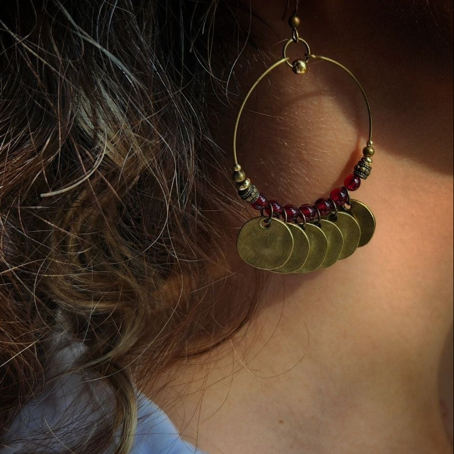 A close-up of a bohemian-style brass hoop earring. The antique brass hoop is decorated with deep-red beads and several hanging, hammered brass coin charms.