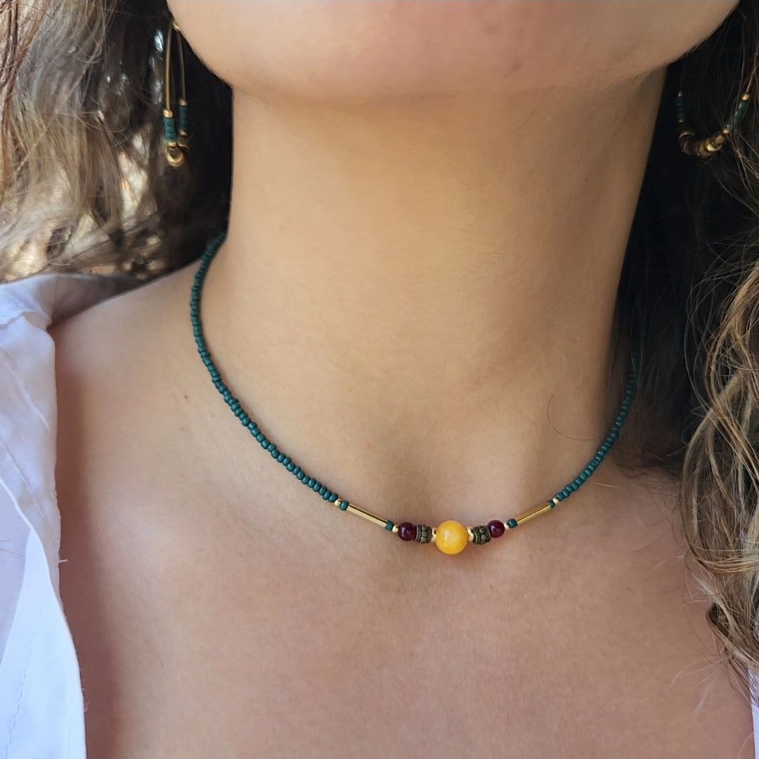 A close-up of a person wearing a delicate, handmade beaded necklace. The necklace consists of small teal seed beads with a central focal arrangement featuring a large yellow bead flanked by gold plated brass spacers, deep-red beads, and gold-tone bar accents.