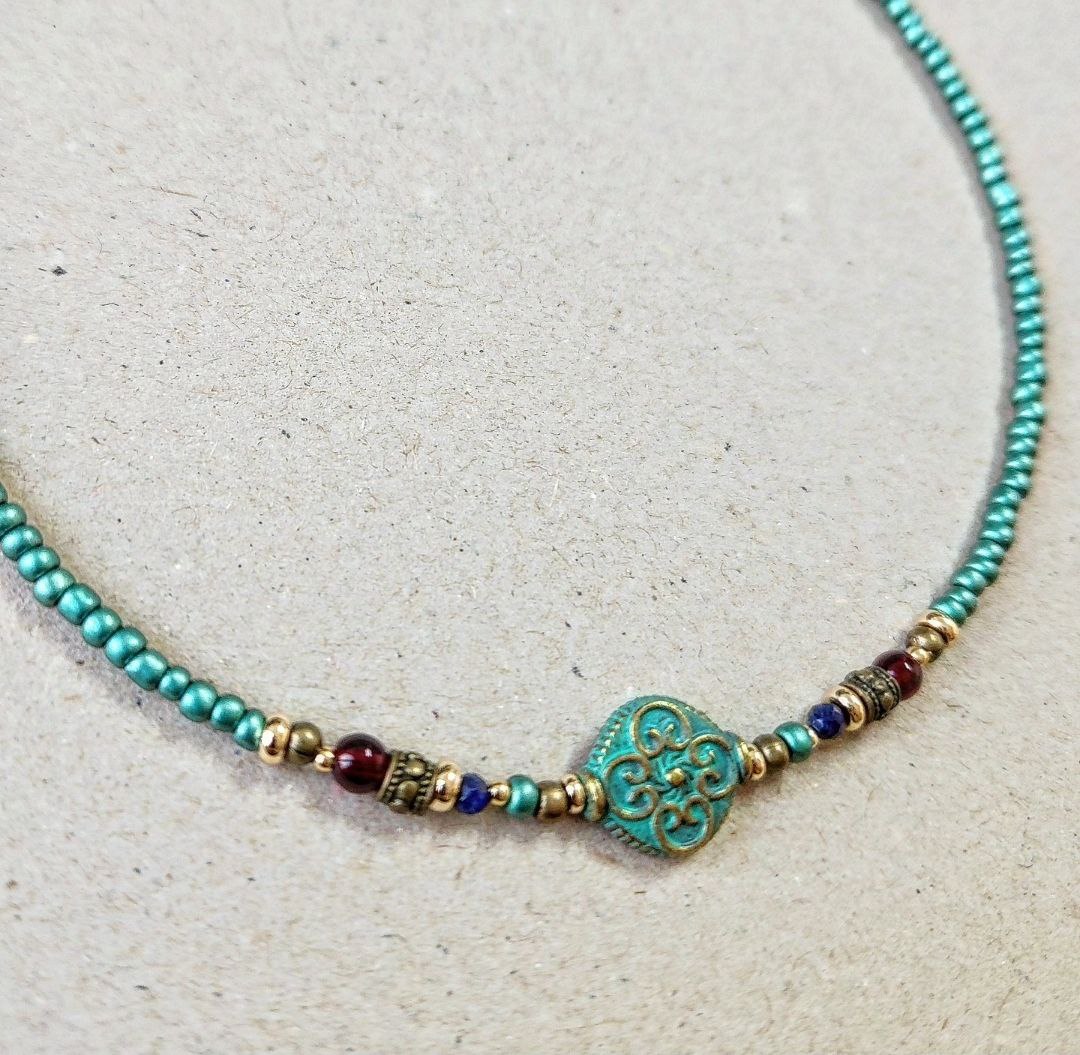 A handmade teal beaded necklace laid flat on a neutral surface. The center features a round patina-green bead with an ornate scroll pattern, flanked by small red, blue, and gold brass decorative beads.