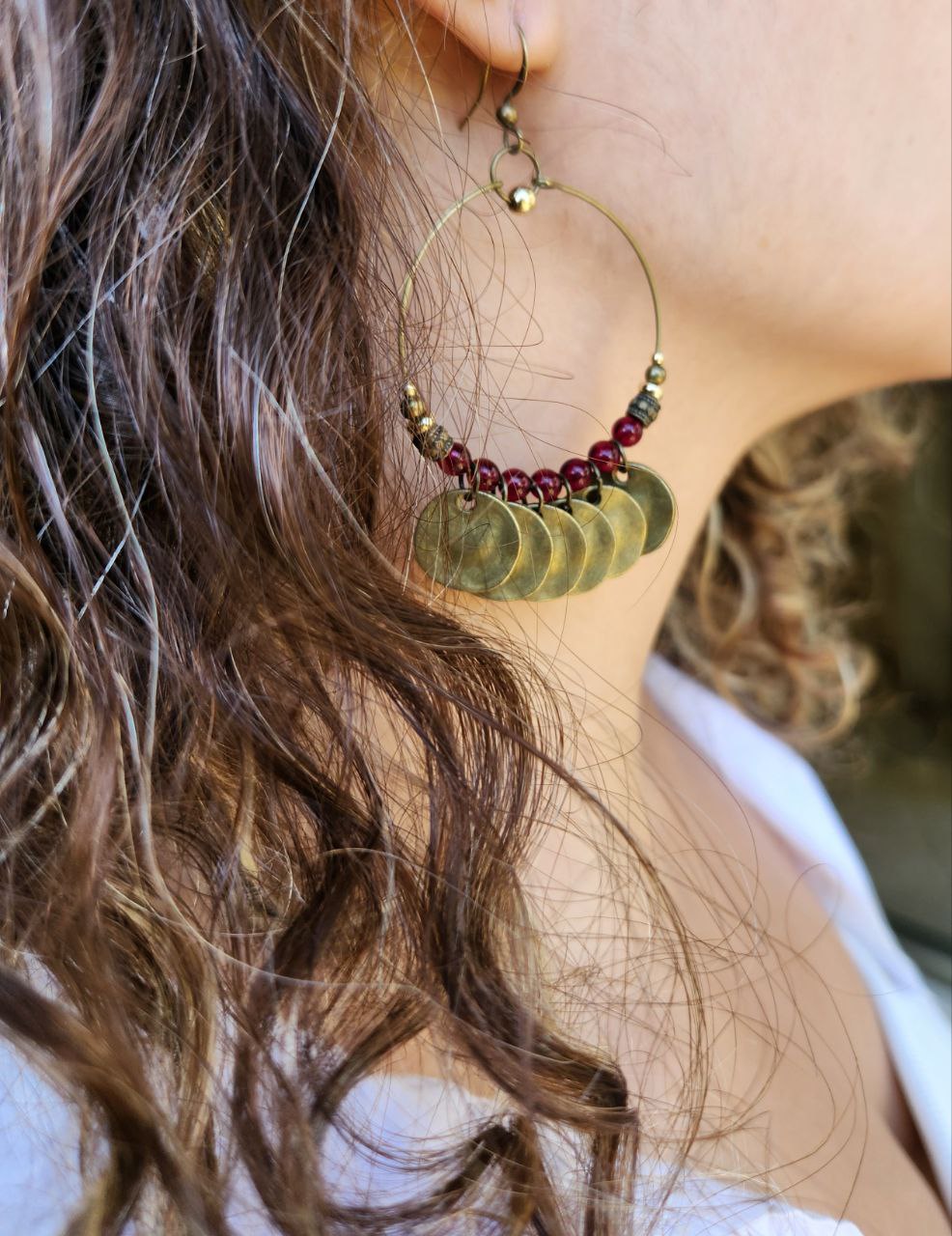 A close-up of a bohemian-style brass hoop earring. The antique brass hoop is decorated with deep-red beads and several hanging, hammered brass coin charms.