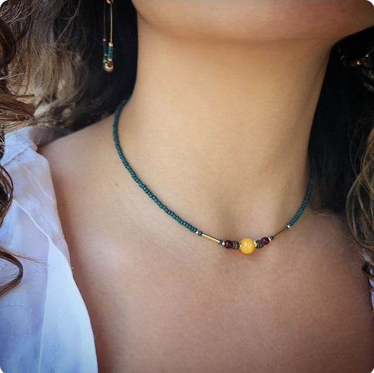 A close-up of a person wearing a delicate, handmade beaded necklace. The necklace consists of small teal seed beads with a central focal arrangement featuring a large yellow bead flanked by gold plated brass spacers, deep-red beads, and gold-tone bar accents.