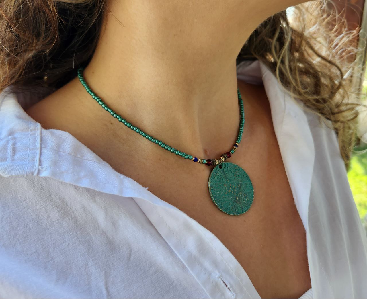 A person wearing a bohemian-style necklace featuring a strand of shimmering teal beads. The centerpiece is a large, circular patina-green pendant attached to the strand with small red and gold plated accent beads.