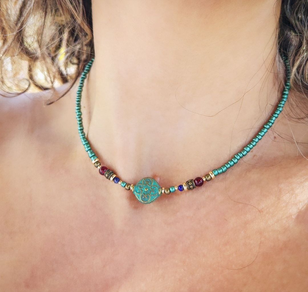 A handmade teal beaded necklace laid flat on a person's neck. The center features a round patina-green bead with an ornate scroll pattern, flanked by small red, blue, and gold brass decorative beads.